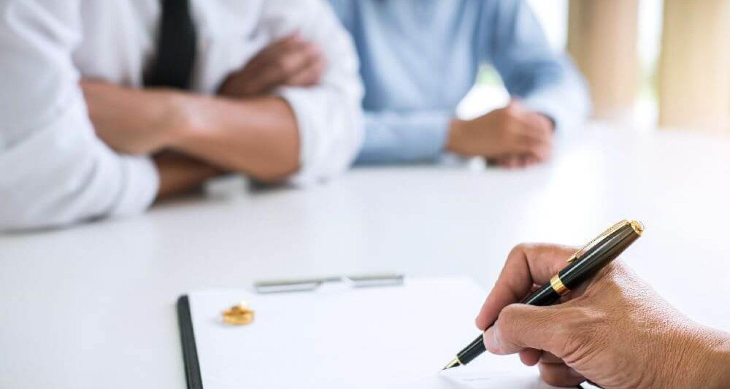 A hand holding a pen poised over a clipboard. Two people with folded arms sit across the table in a bright, neutral meeting room.