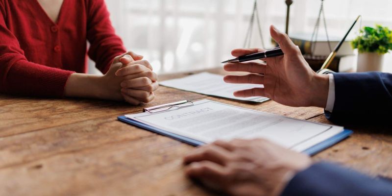 Person in a red sweater sits at a wooden table with clasped hands as another person in a suit offers a pen over a contract on a clipboard.