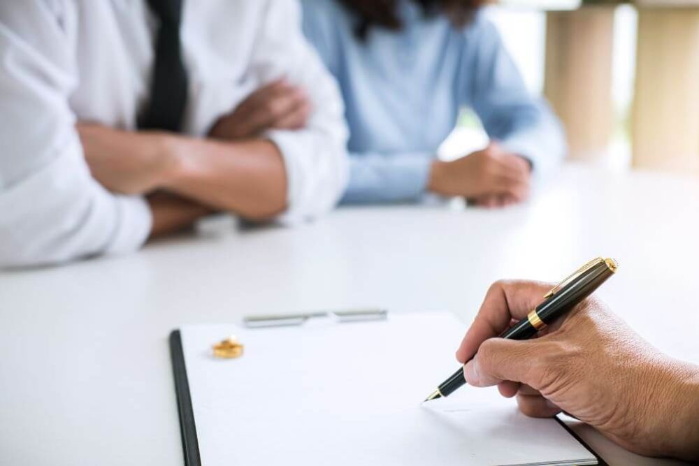 A hand holding a pen poised over a clipboard. Two people with folded arms sit across the table in a bright, neutral meeting room.