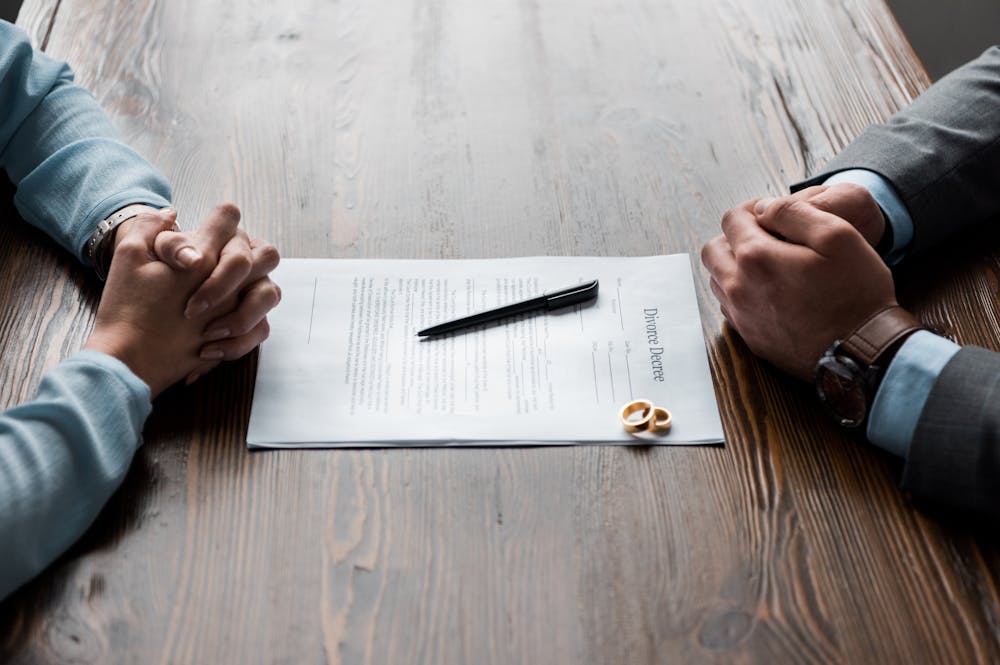Two pairs of clasped hands rest on a wooden table, facing each other over a divorce decree document with a pen and two wedding rings placed on it.